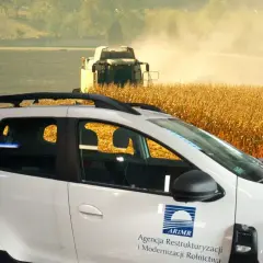 Biały samochód z logo ARiMR na tle żniw kukurydzy. Nowoczesne rolnictwo, które kiedyś było kolektywizacją, dziś wygląda tak.