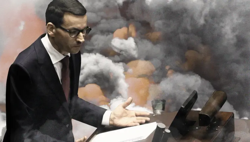 Mateusz Morawiecki w garniturze przemawia na tle dymu i pomarańczowych płomieni.